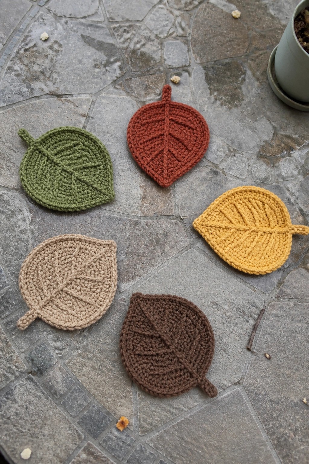 Five colorful knitted leaf coasters in autumn hues on gray stone pavement.