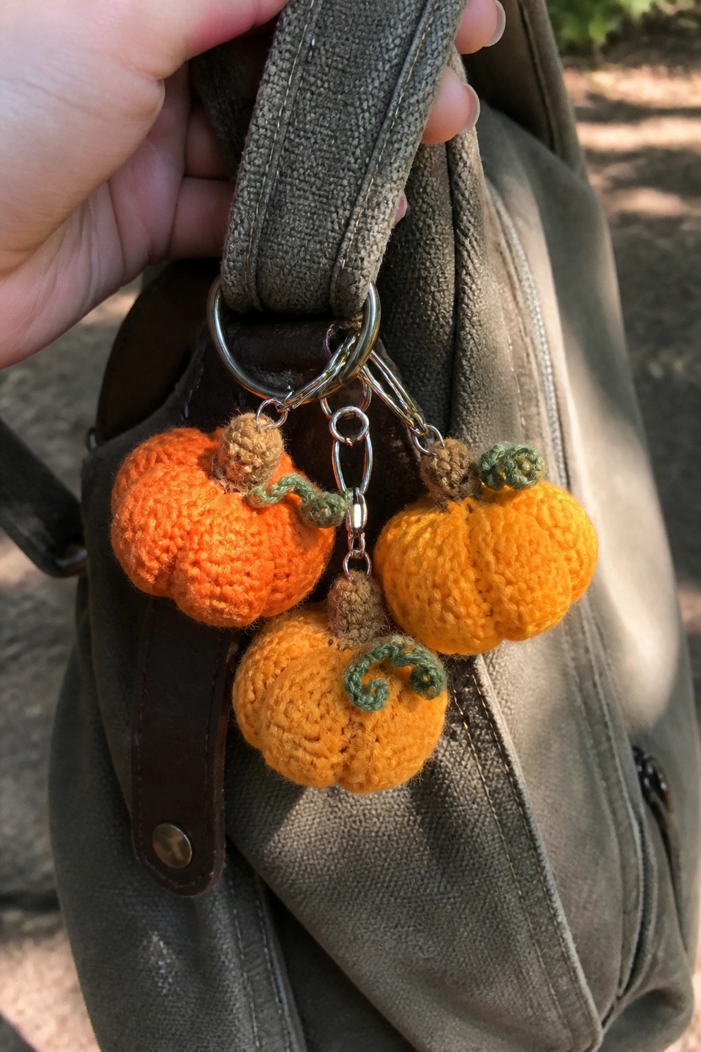 Hand holding gray bag with three crocheted orange pumpkin keychains on metal clips