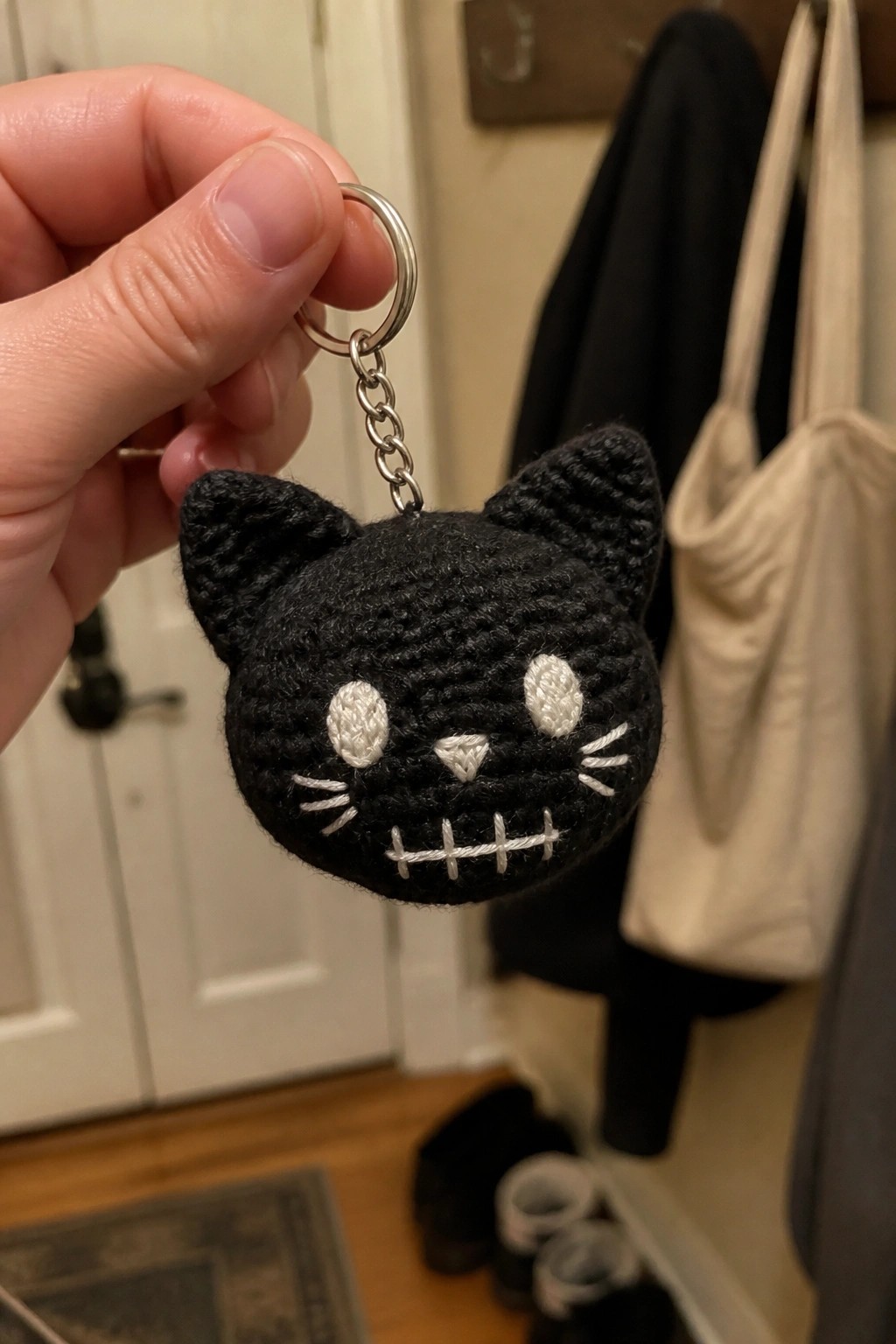 Hand holding black crocheted cat keychain with white stitched facial features.