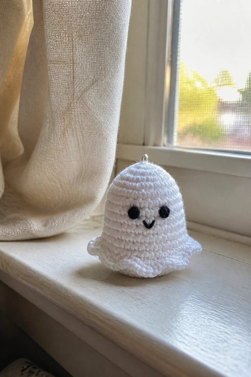 Small white crocheted ghost with black eyes on sunny windowsill beside curtain.