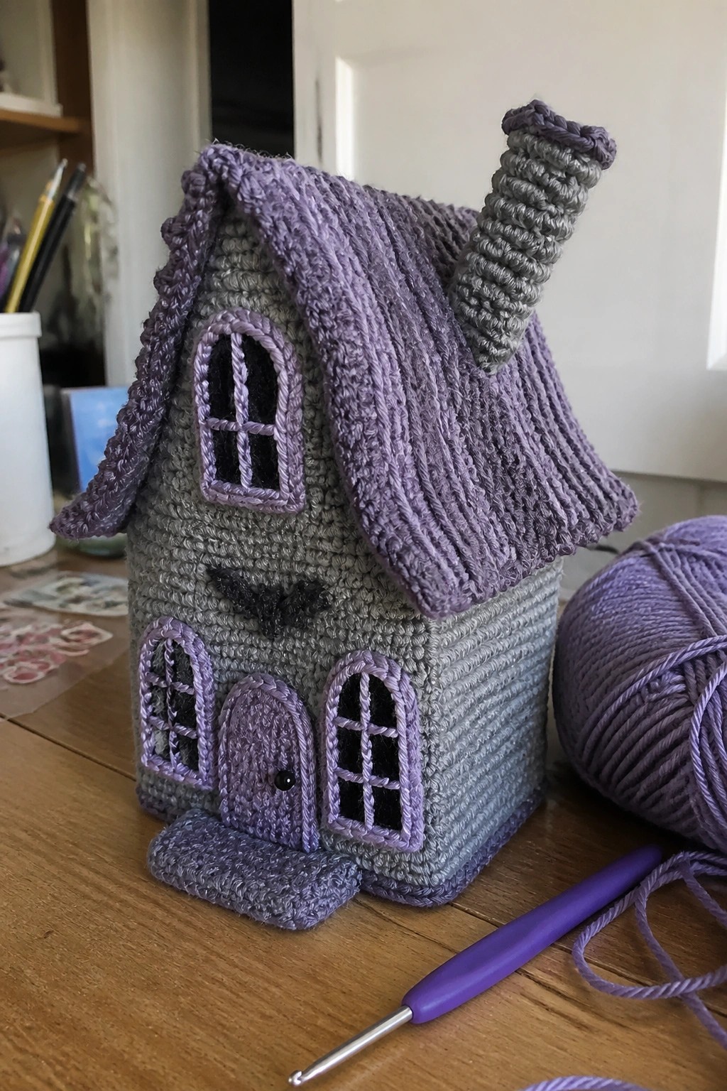 Crocheted purple-gray house with chimney, windows, door, yarn ball, and hook.