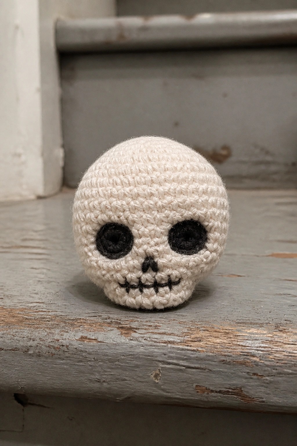 A small white crocheted skull with black embroidered eyes, nose, and mouth sits on a wooden surface.
