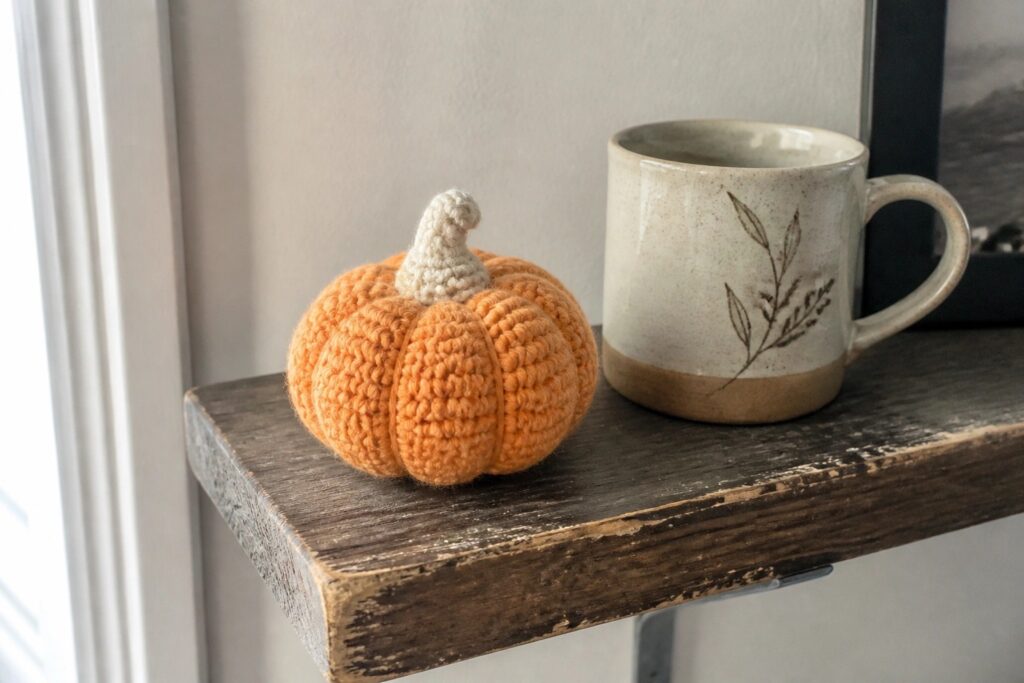 A small crocheted pumpkin with orange and cream stripes and a beige stem sits on a wooden shelf next to a ceramic mug.