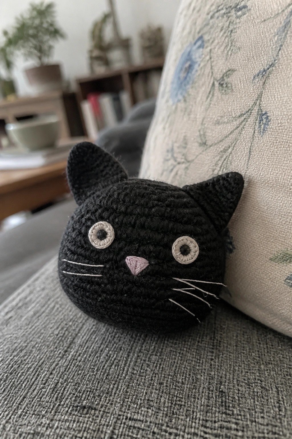 Black crocheted cat head with pink nose and white eyes on gray couch beside floral pillow