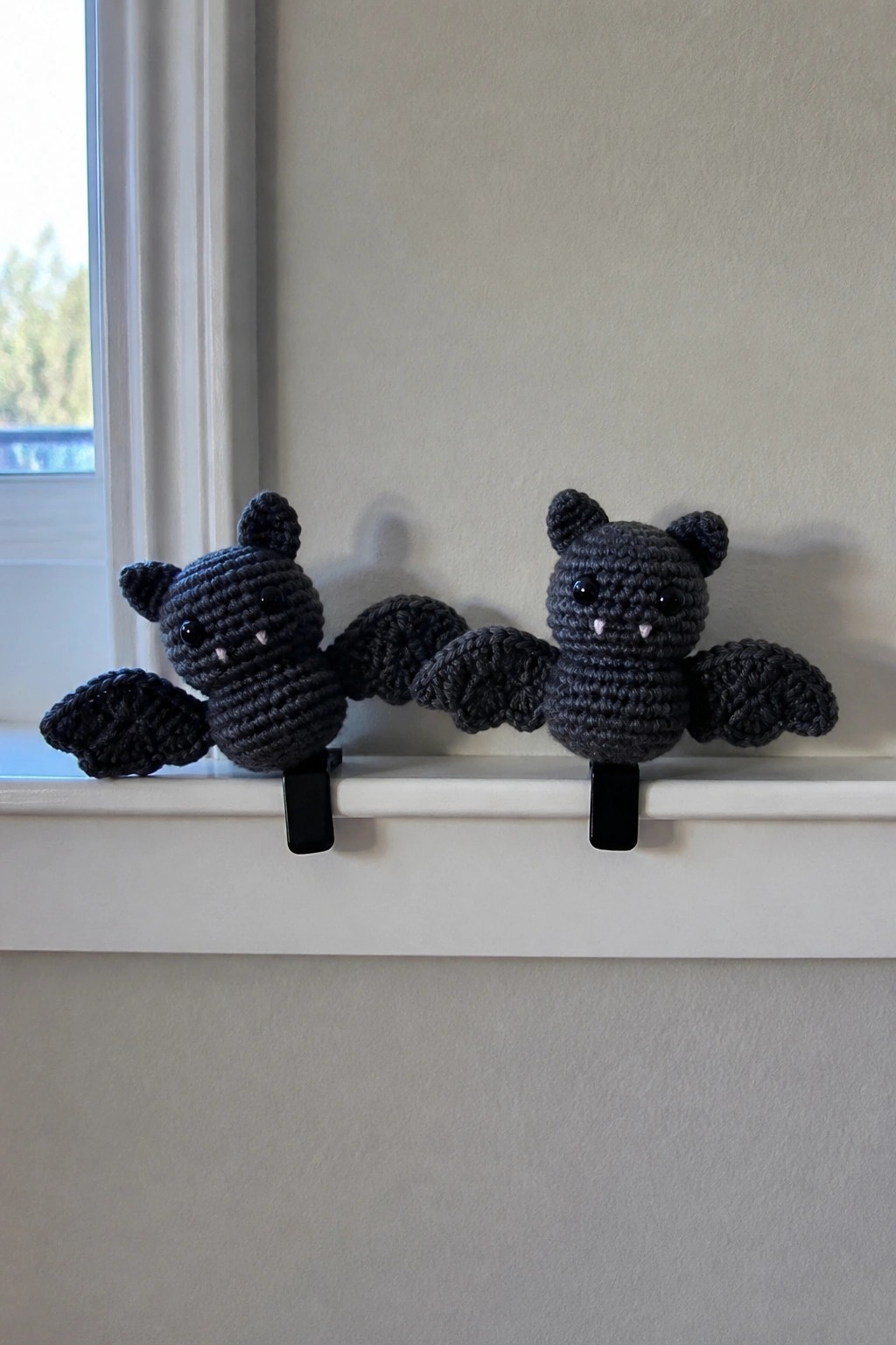 Two crocheted black bats clipped to a white windowsill beside a window.