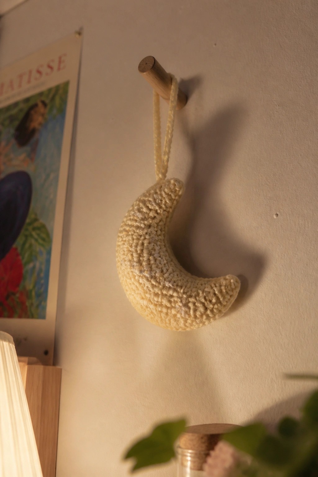 Crocheted cream crescent moon hanging from wooden peg on wall near poster