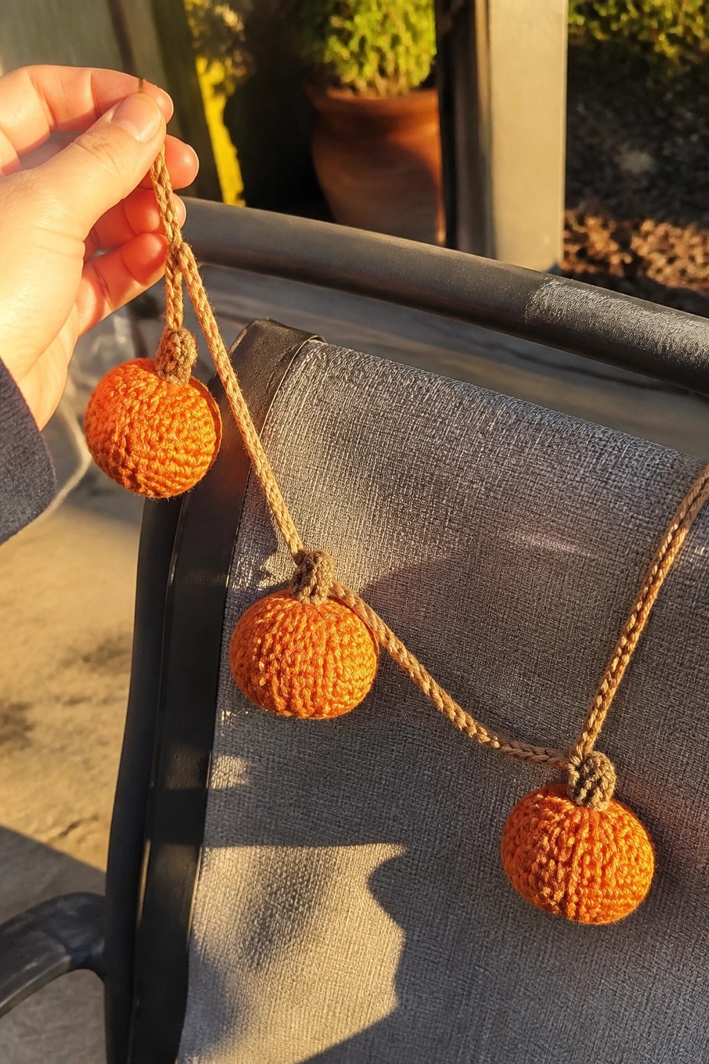 Hand holding three orange crocheted pumpkins strung on brown twine in sunlight.