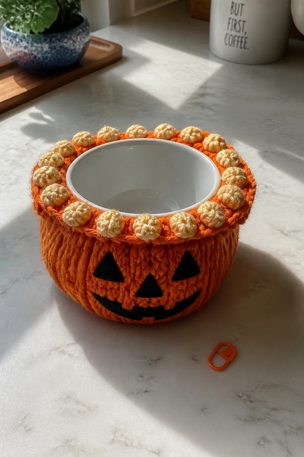 Crocheted orange pumpkin jack-o'-lantern cozy around white bowl on marble counter