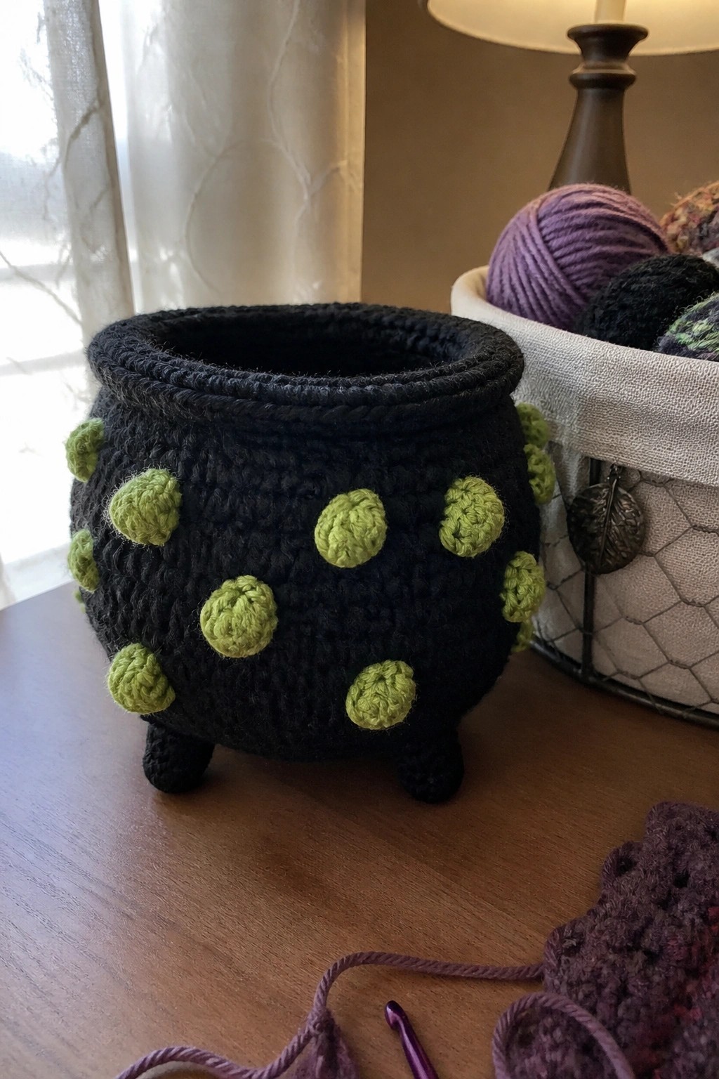Black crocheted cauldron with green bobbles on wooden table near yarn and hook