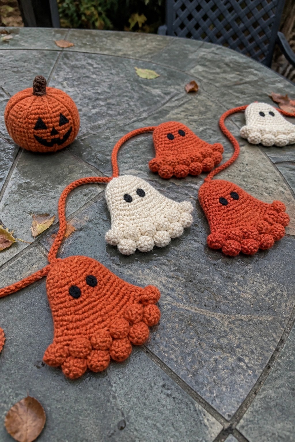 Crocheted orange pumpkin and white ghost garland on stone table with leaves