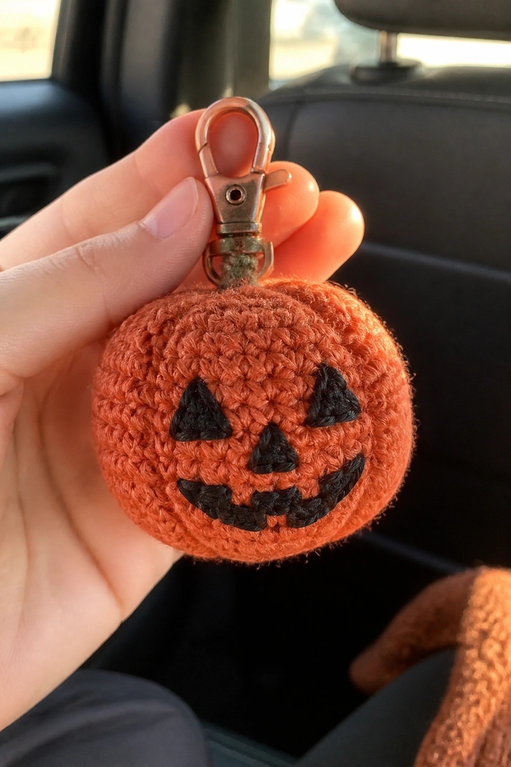 Hand holding orange crocheted pumpkin keychain with black jack-o'-lantern face and metal clasp.