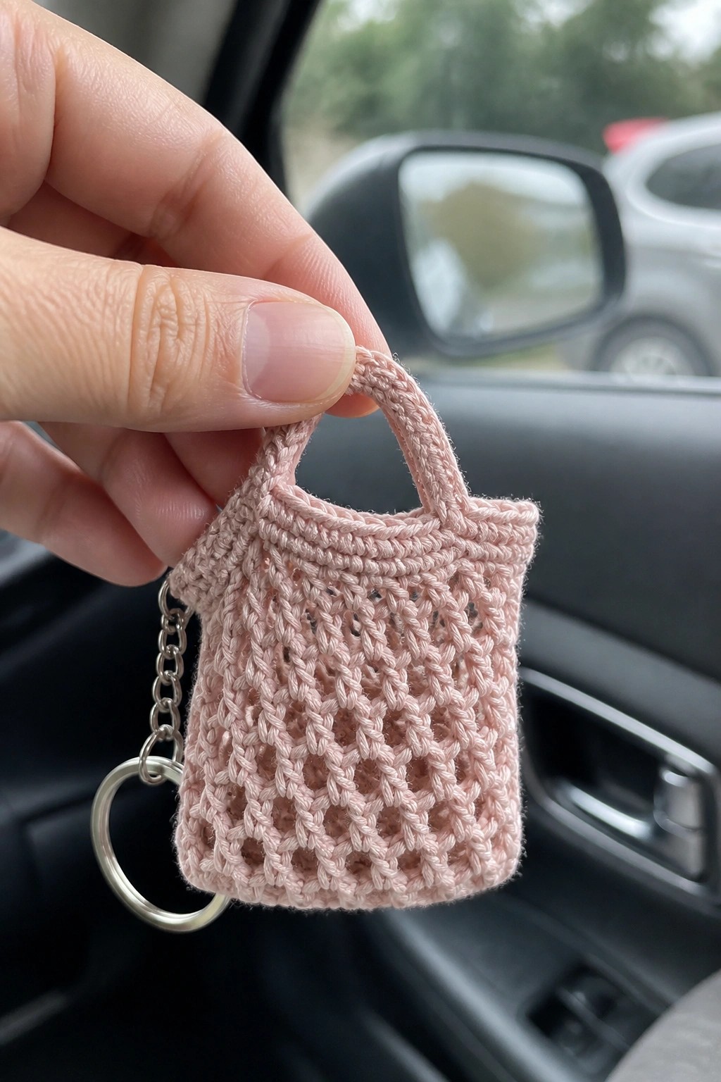 Hand holding small pink crocheted tote bag keychain in car interior