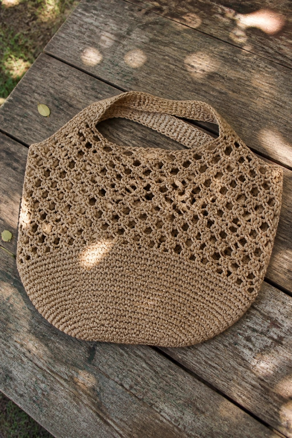 Beige crocheted mesh tote bag on sunlit weathered wooden table.