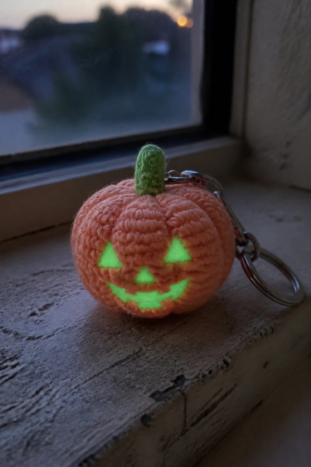 Crocheted orange pumpkin keychain with glowing green jack-o'-lantern face on windowsill
