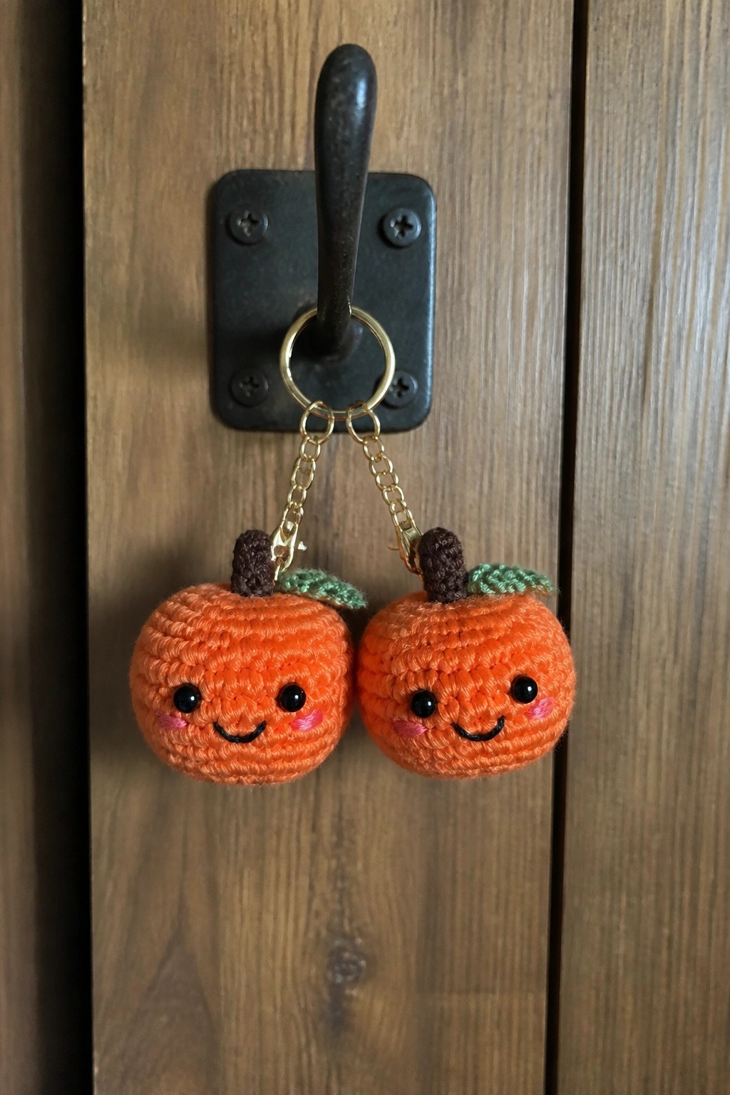 Two crocheted orange pumpkin keychains with faces hang from a black hook on wood.