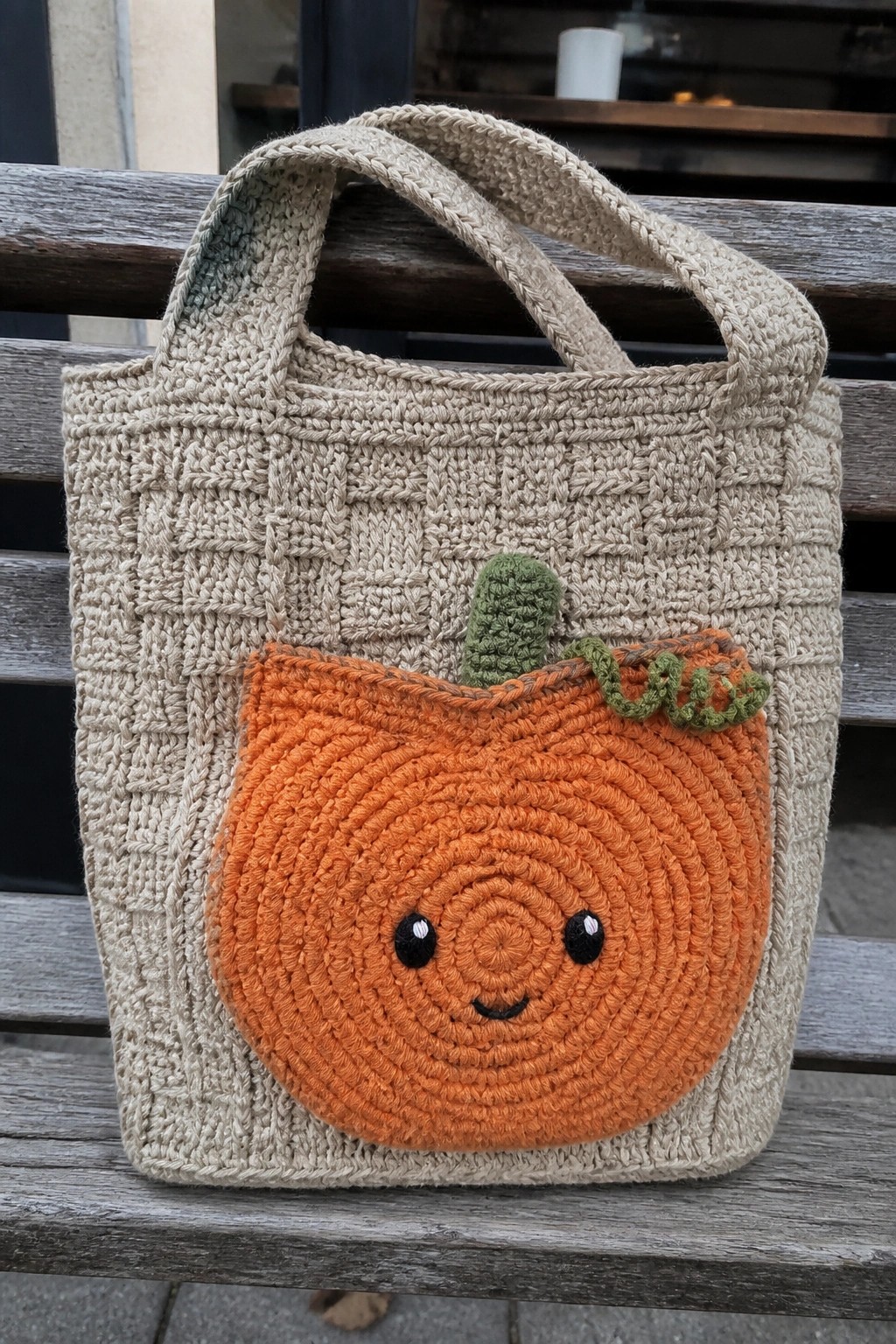 Beige crocheted tote bag with smiling orange pumpkin face and green stem on wooden bench.