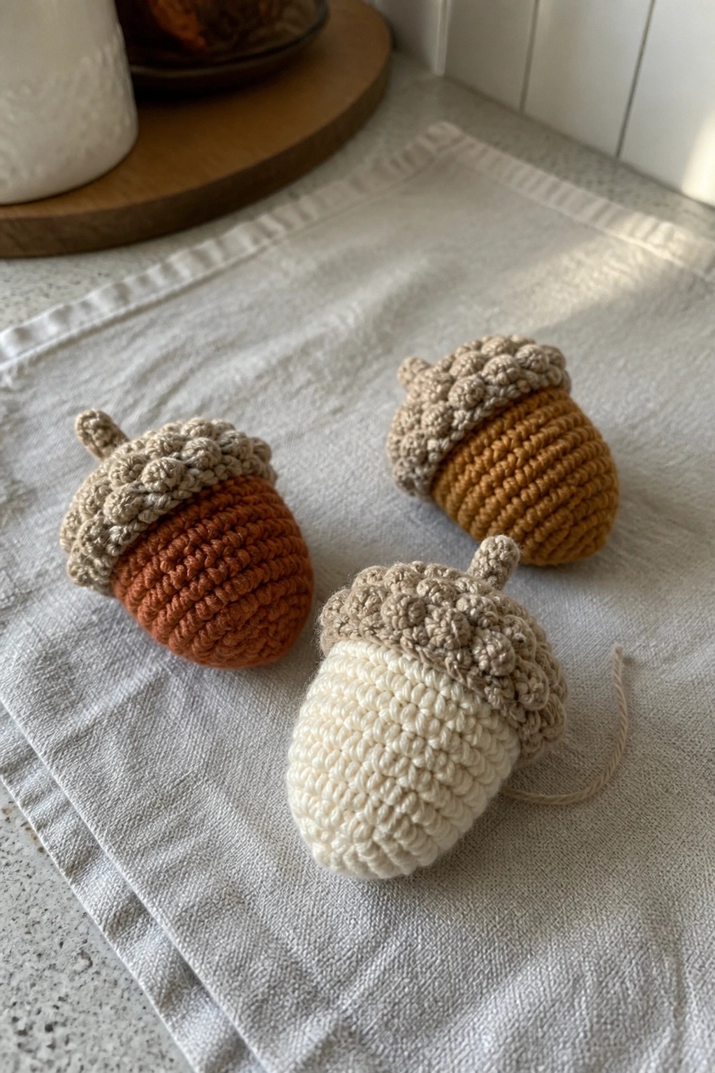 Three small crocheted acorns in rust, mustard, and cream rest on a linen cloth.