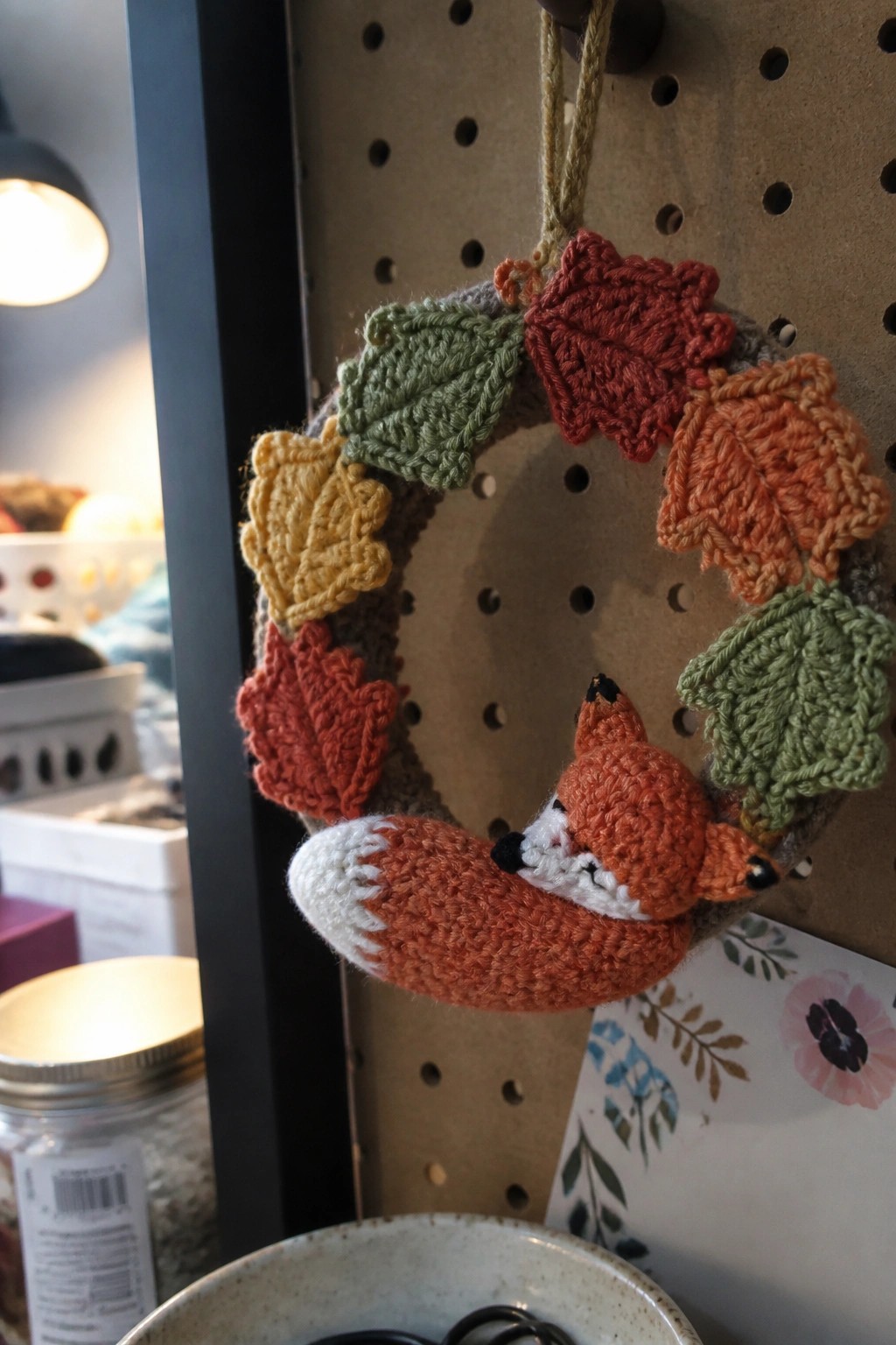 A crocheted autumn wreath with multicolored leaves and a small fox figure hangs from a peg on a pegboard.