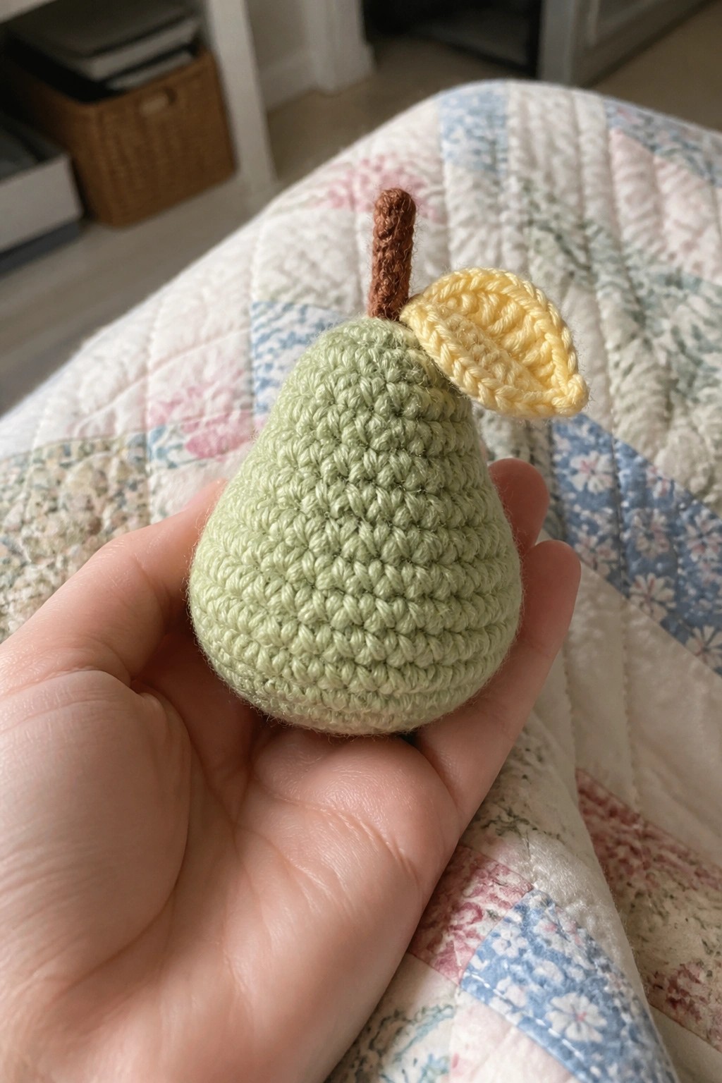 Hand holding crocheted green pear with yellow leaf and brown stem on floral quilt.
