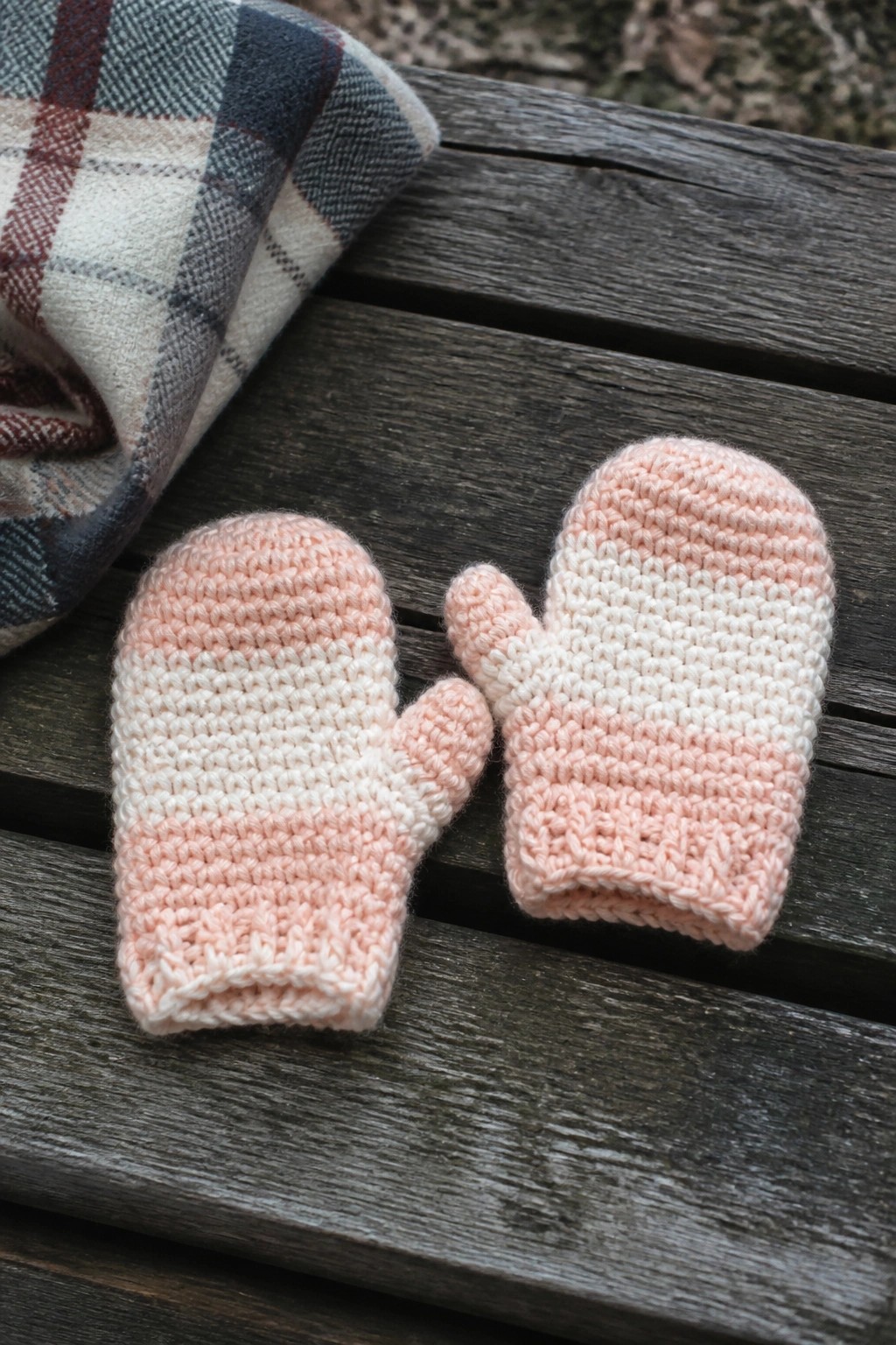 Two crocheted mittens with pink and white stripes sit on a wooden surface beside a plaid blanket.