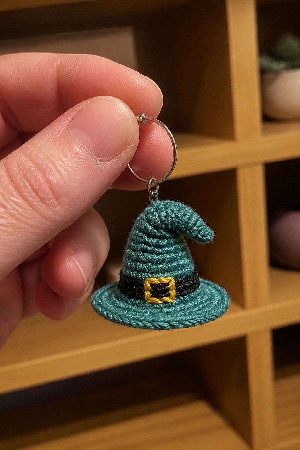 Hand holding miniature teal crocheted witch hat keychain with yellow buckle.