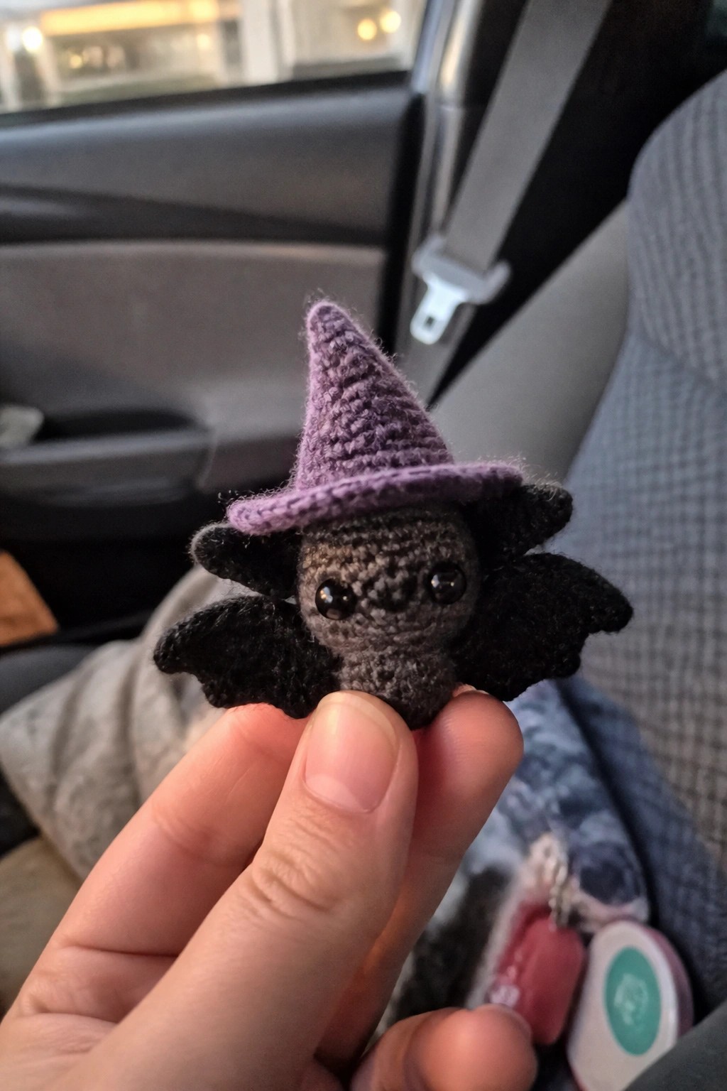 Tiny gray crocheted bat wearing purple witch hat held by fingers in car.