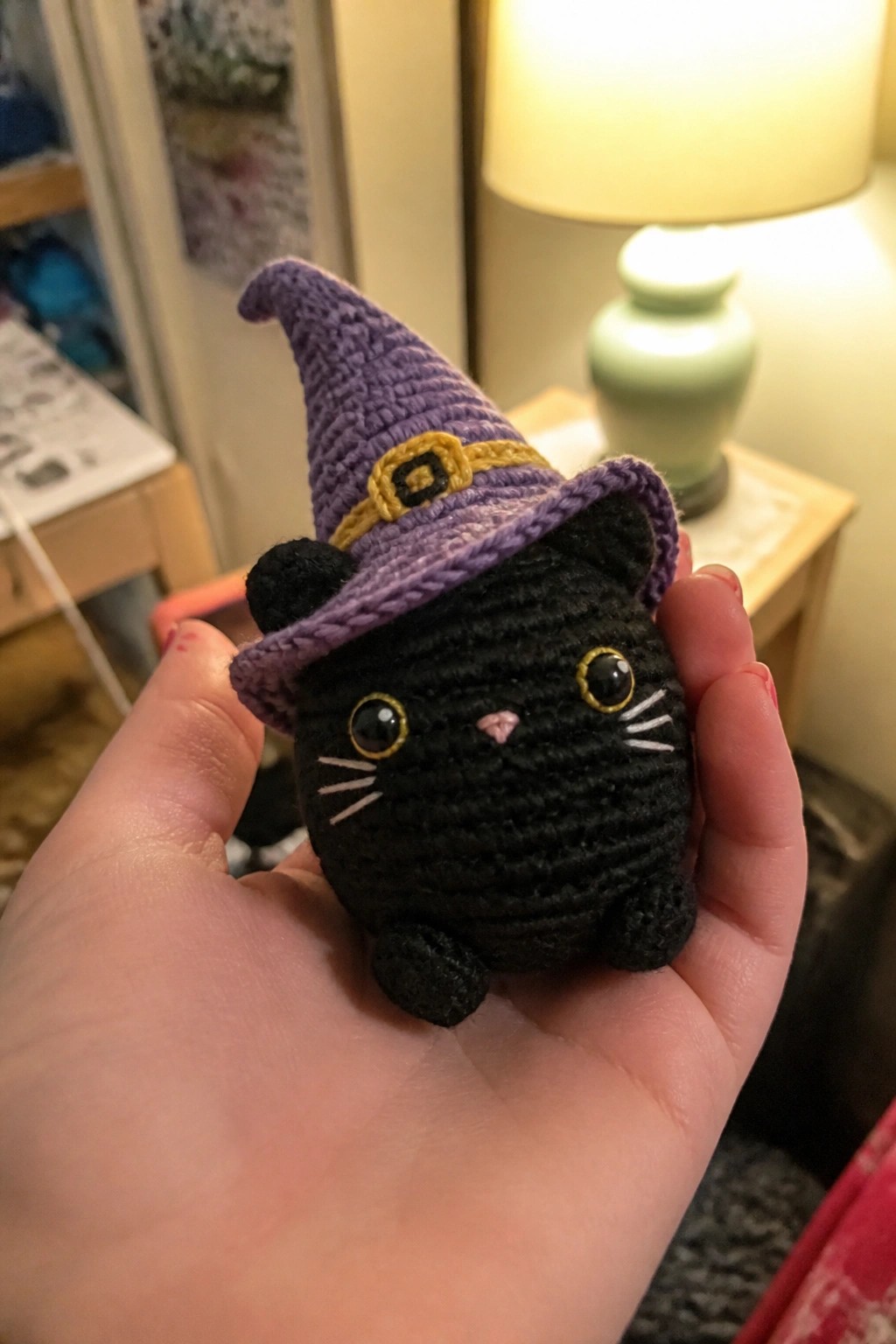 Hand holding small black crocheted cat wearing purple witch hat with gold buckle.