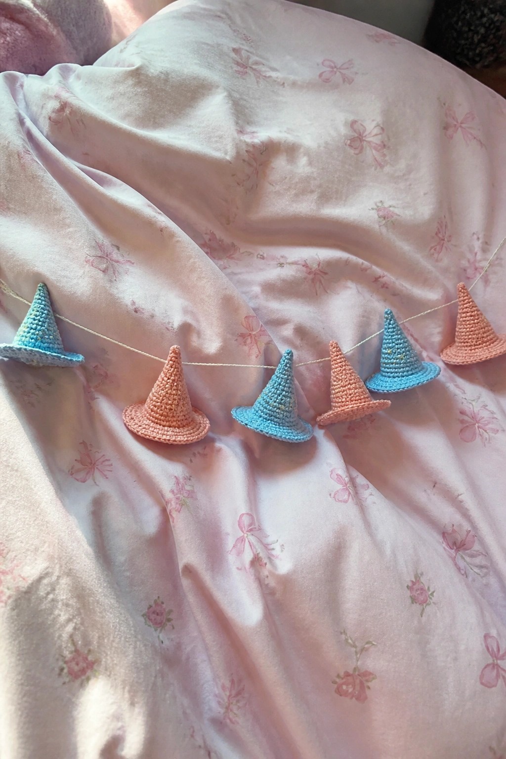 Pastel crocheted witch hats strung across pink floral fabric.