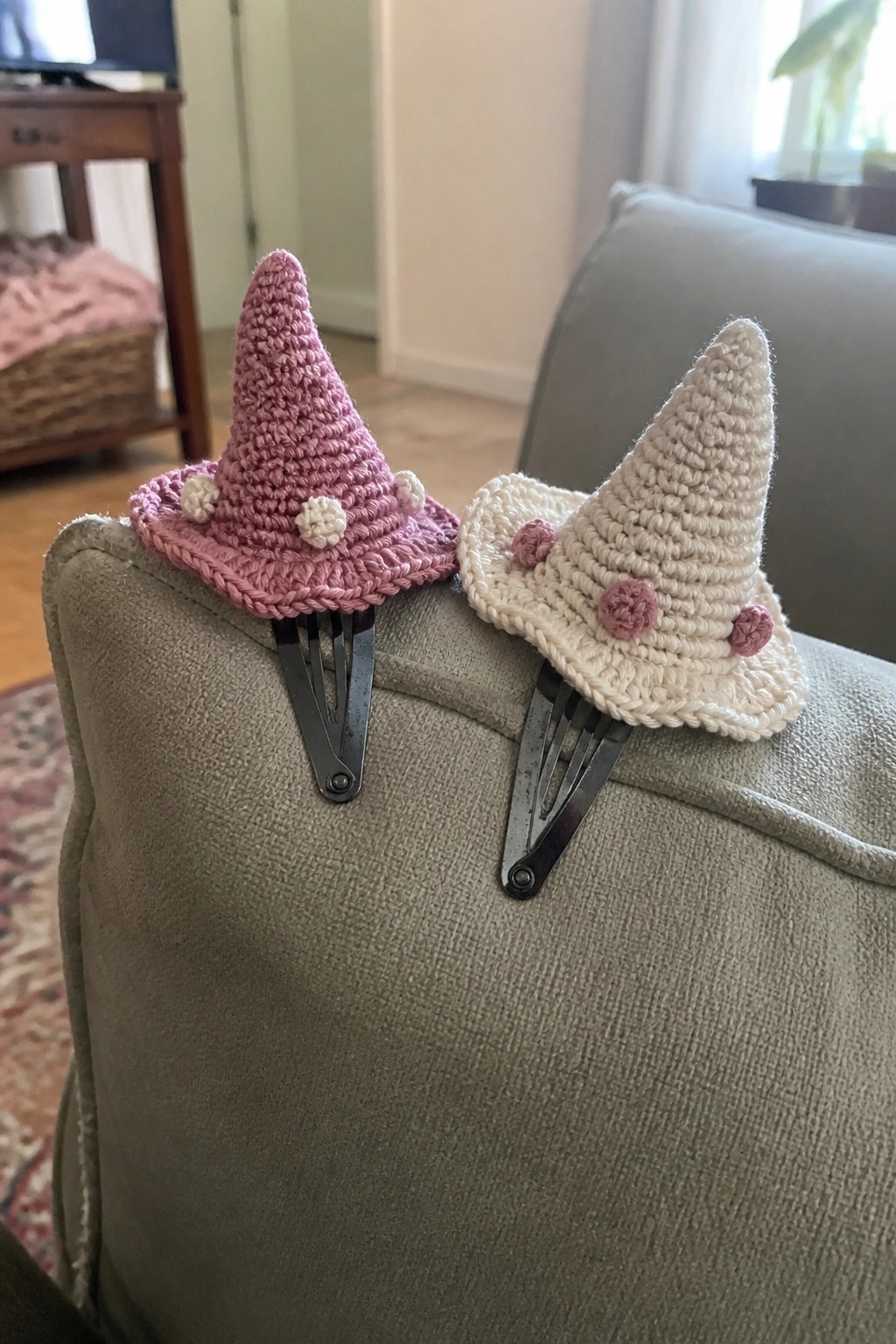 Pink and cream crocheted witch hats with pom-poms on hair clips atop gray couch