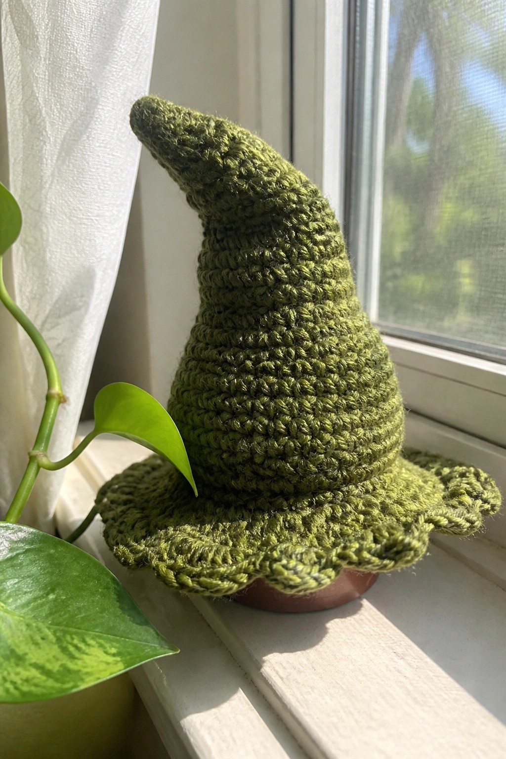 Green crocheted witch hat with scalloped brim on sunny windowsill plant pot