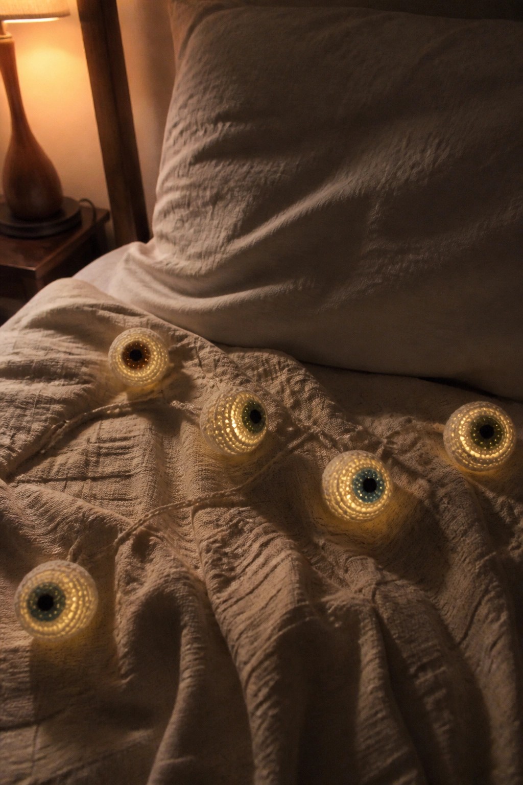 Glowing eyeball string lights scattered on rumpled white bedsheets in a cozy bedroom.