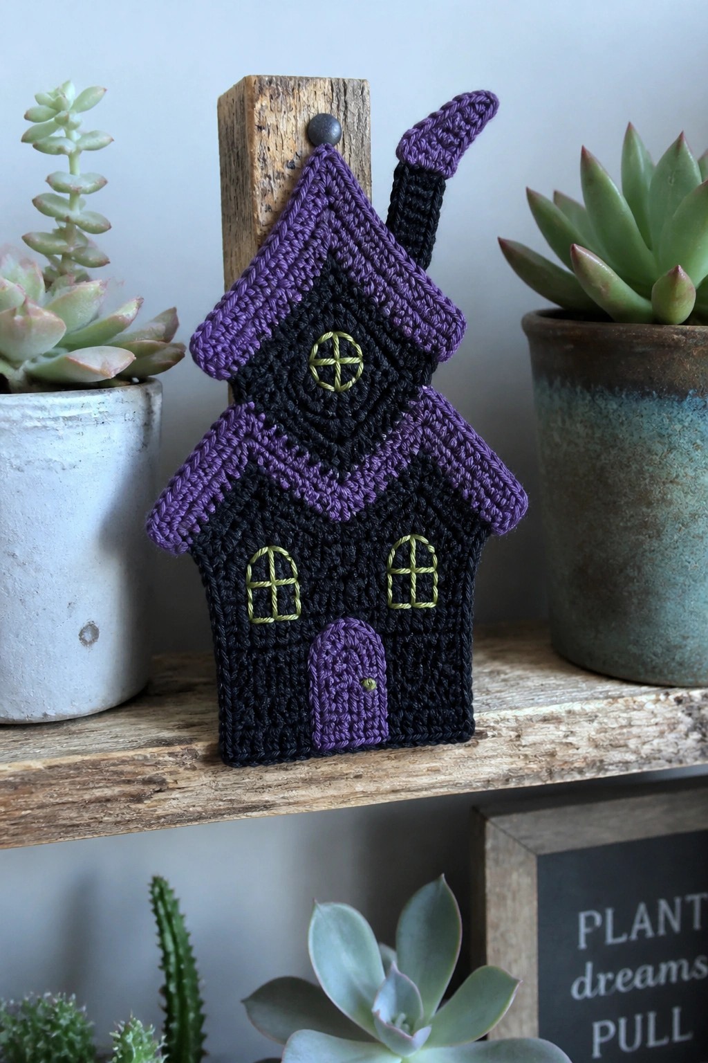 Crocheted black and purple house ornament hanging among potted succulents on wooden shelf