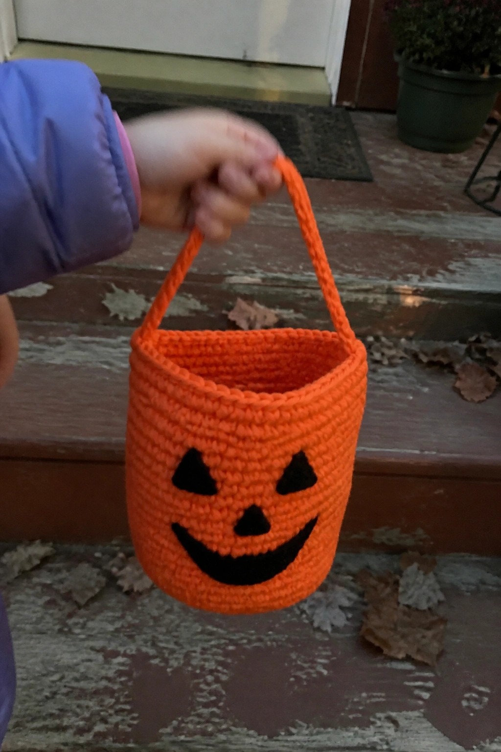 Child's hand in blue jacket holding orange crocheted jack-o'-lantern bucket on steps