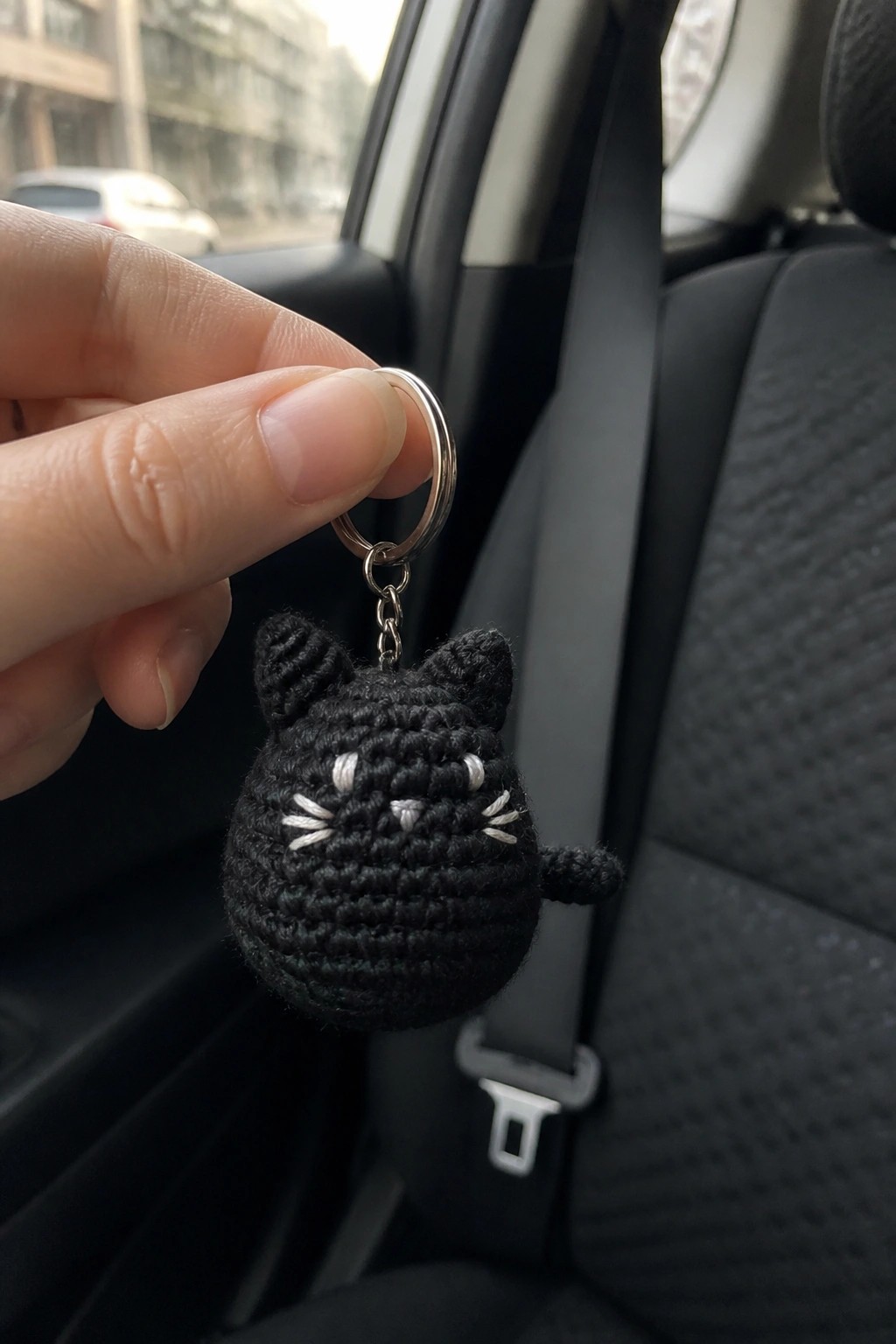 Hand holding black crocheted cat keychain with white whiskers in car seat