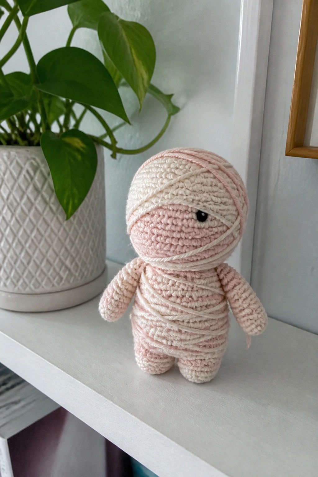 A small crocheted mummy figure in pink and white yarn sits on a white shelf next to a potted plant.