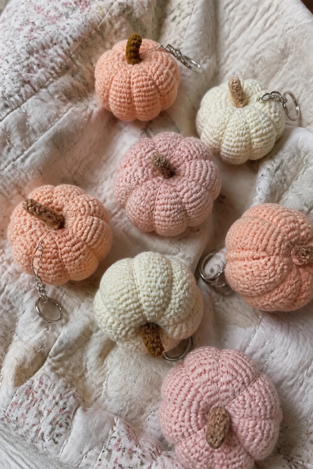Crocheted pumpkin keychains in pastel pink, peach, and white on floral quilt.