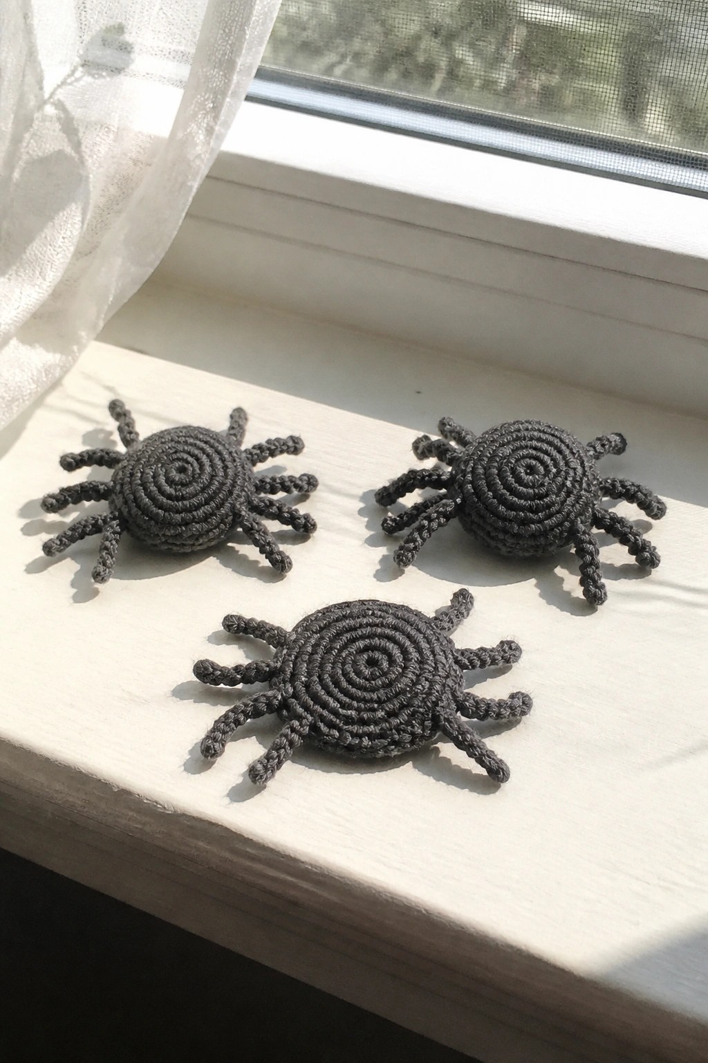 Three dark gray crocheted spiders on a sunlit white windowsill