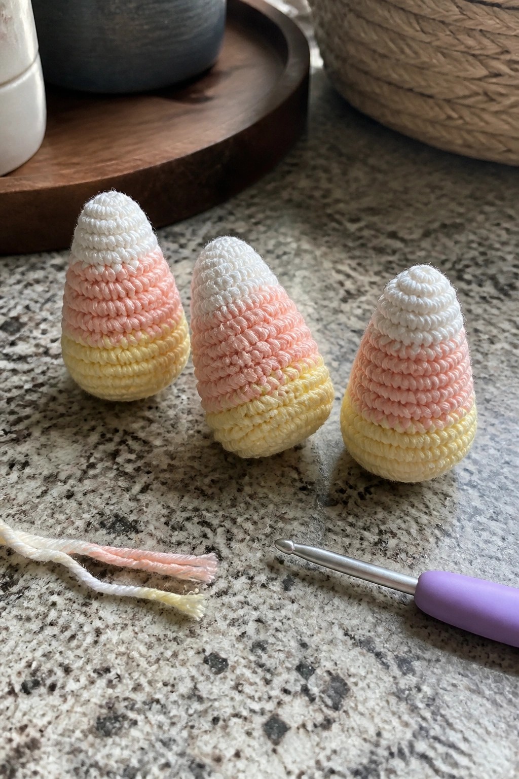 Three crocheted candy corns in white, pink, and yellow with hook and yarn