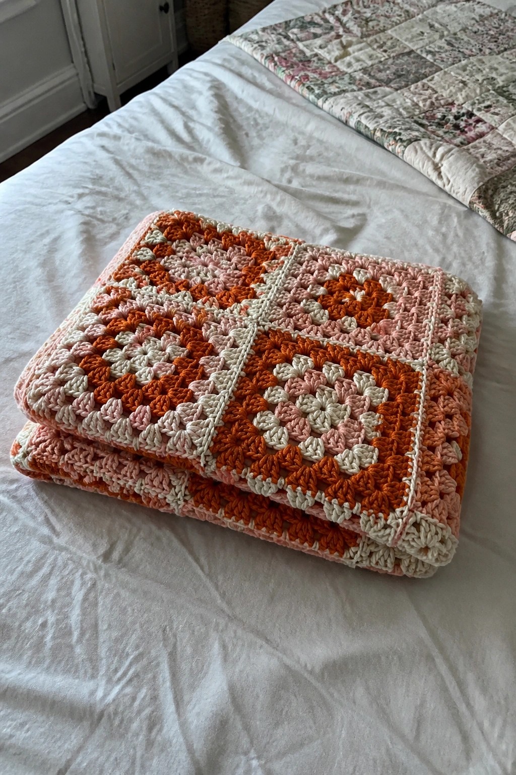 Folded orange and pink crocheted granny square blanket on white bed with quilt