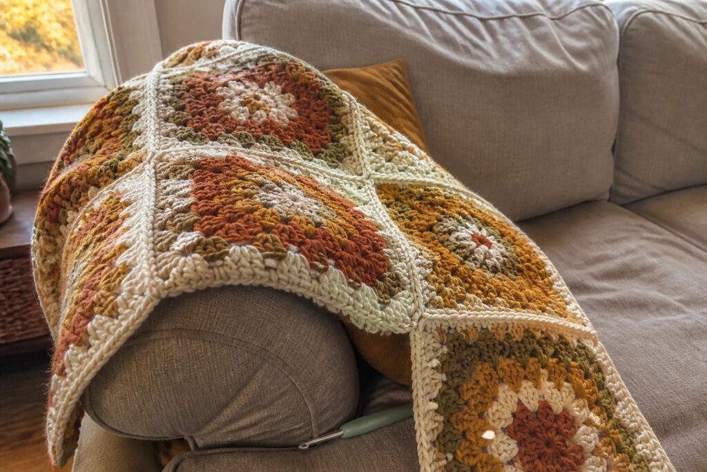 25 Cozy Fall Crochet Blanket Ideas Featuring Autumn Granny Squares A crocheted blanket of autumn-colored granny squares lies across a gray couch with a crochet hook nearby.