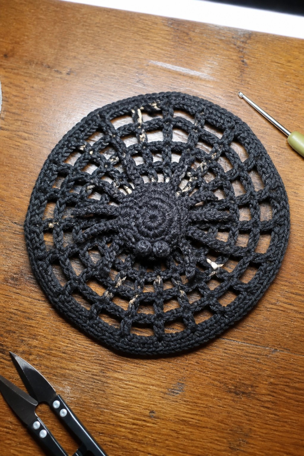 Black crocheted doily with lace pattern on wood beside hook and scissors