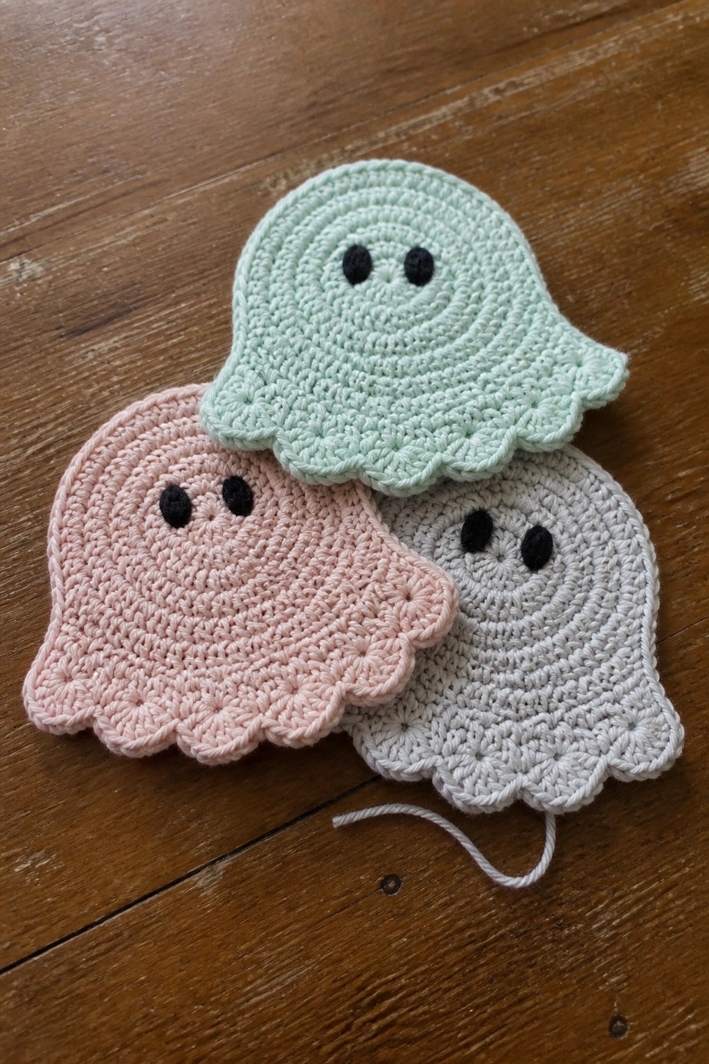 Three pastel crocheted ghosts in green, pink, and gray on wooden surface