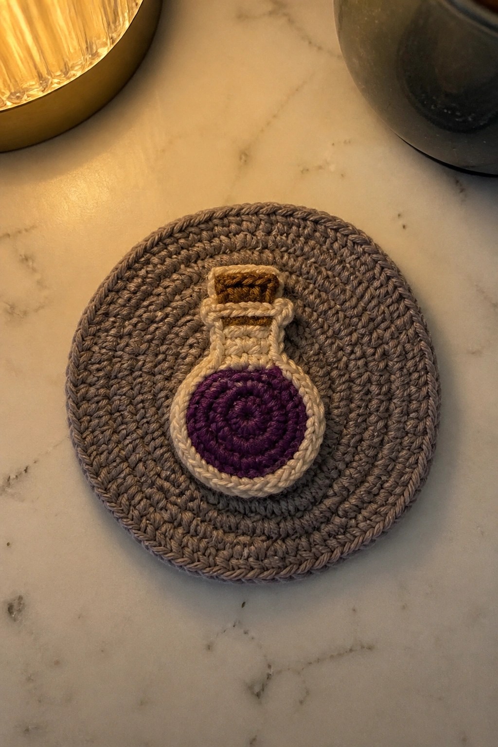 Gray crocheted coaster with purple potion bottle appliqué on marble.