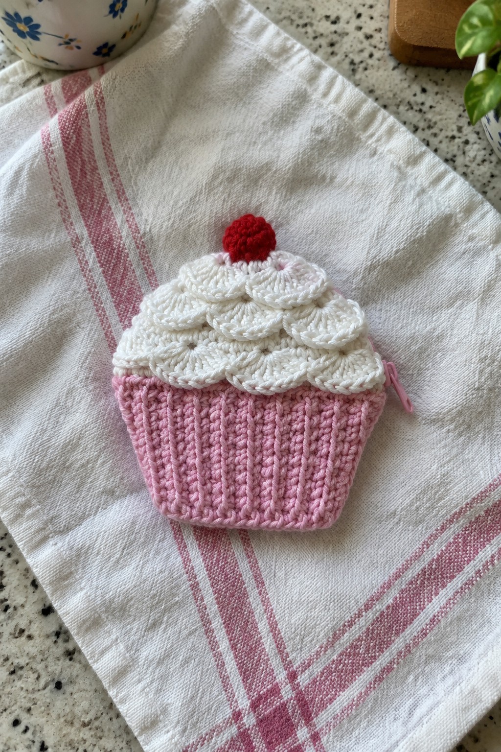 Crocheted pink cupcake pouch with white scalloped top and red cherry on striped towel.