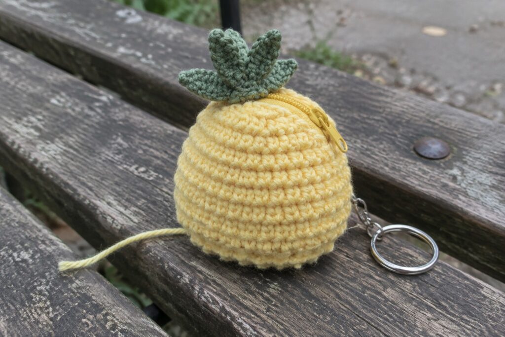 Yellow crocheted pineapple pouch with green top, zipper, and keyring on wooden bench.