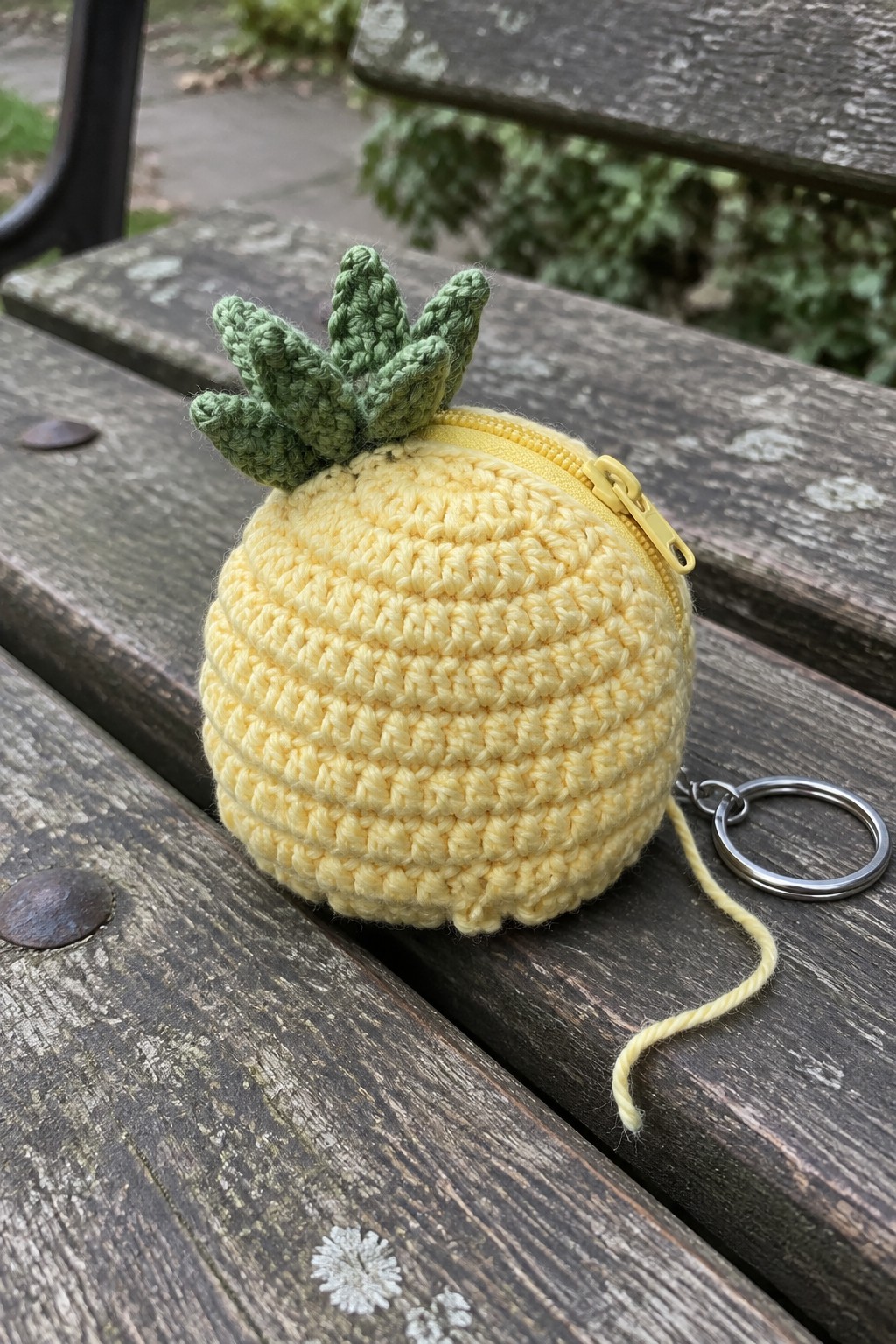 Yellow crocheted pineapple pouch with green top, zipper, and keyring on wooden bench.
