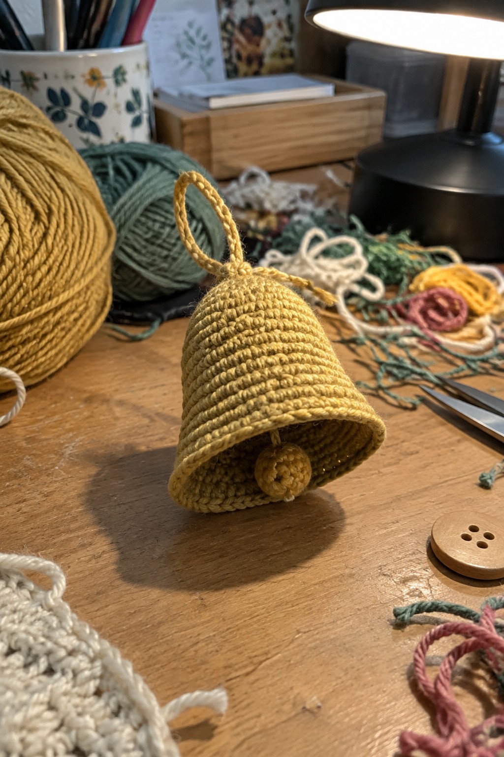 Yellow crocheted bell ornament on wooden table amid yarn balls and supplies