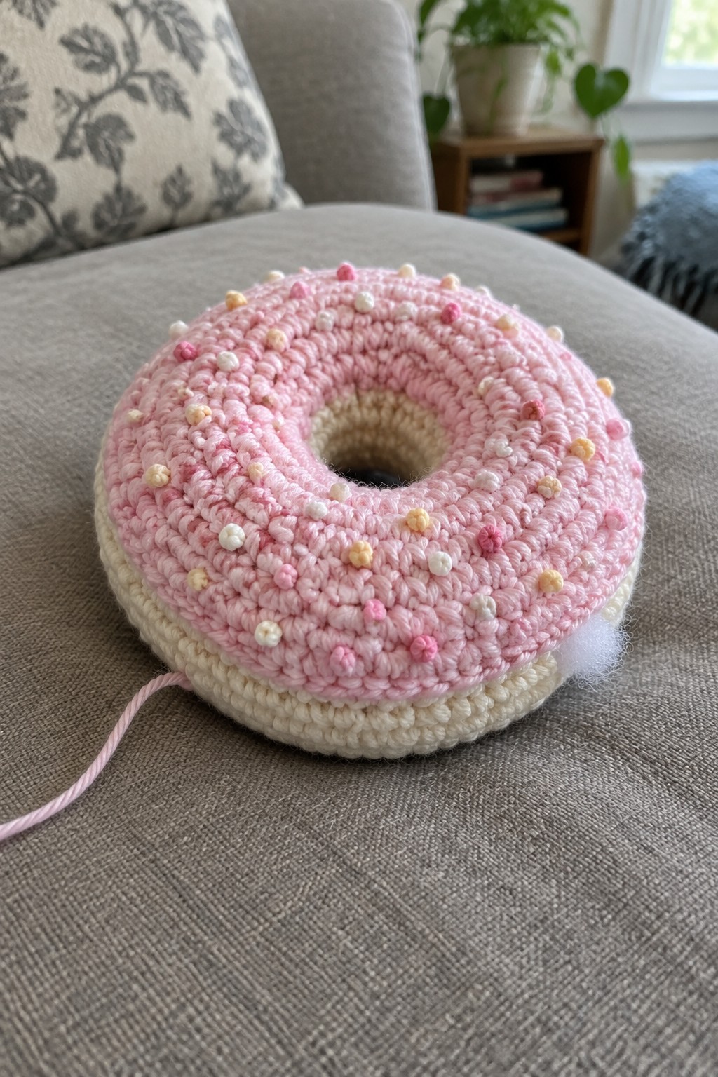 Pink crocheted donut plush with colorful sprinkles on gray couch.