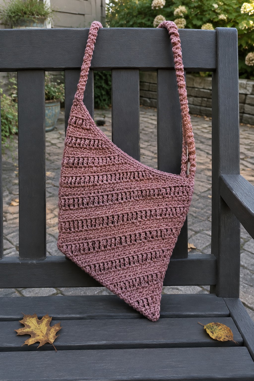 Pink crocheted triangular bag draped over dark wooden bench with autumn leaves