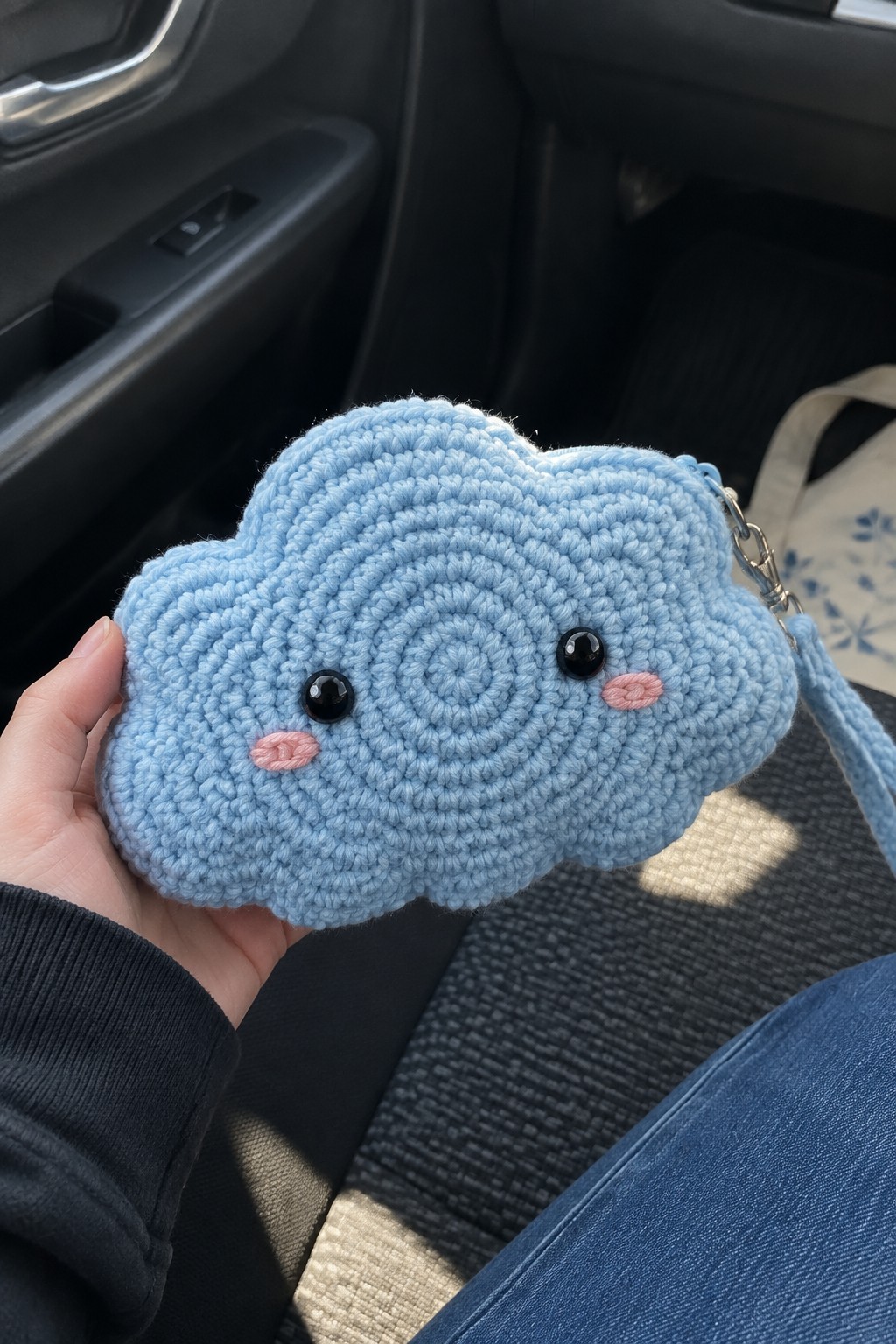 Hand holding light blue crocheted cloud pouch with black eyes and pink cheeks in car.